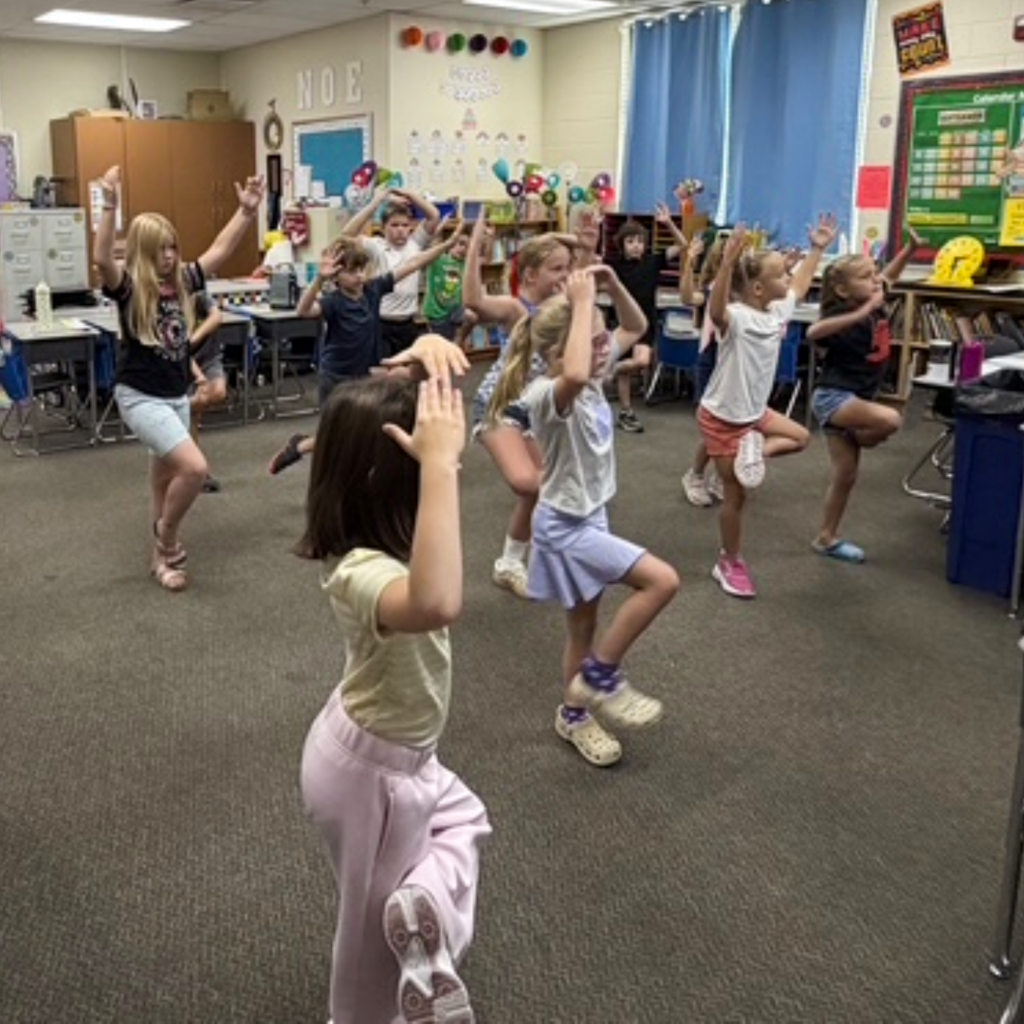 A class of students standing with their hand above their heads in their classroom.