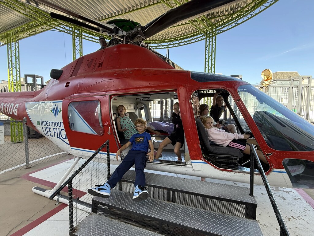Students sitting inside and posing around a life-size red Intermountain Life Flight helicopter exhibit on an outdoor deck platform.