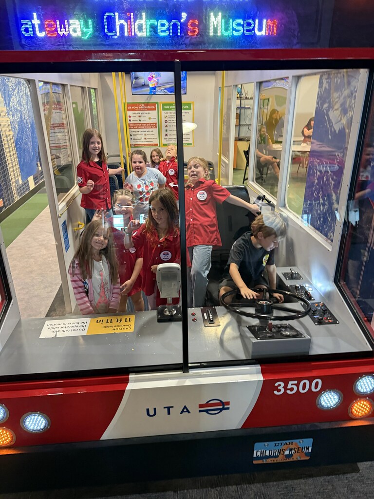 A group of students standing inside a mock Utah Transit Authority (UTA) bus exhibit, with one student sitting in the driver's seat.