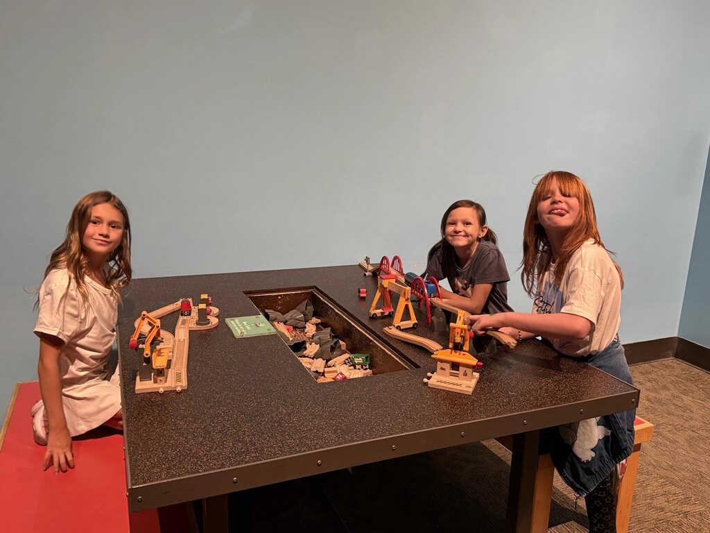 Three students smiling around a wooden train table exhibit filled with wooden tracks, bridges, and toy trains.
