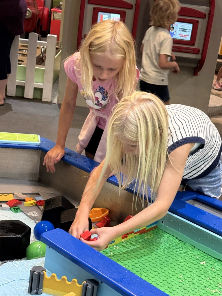 Two girls leaning over a water table exhibit, playing with floating plastic blocks and boat toys.