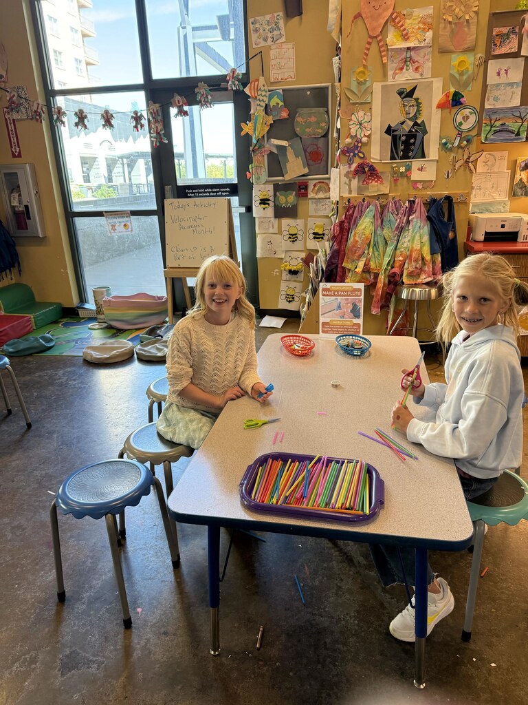 Two girls sitting at an art table filled with colorful craft straws, scissors, and a sign titled "Make a Pan Flute."