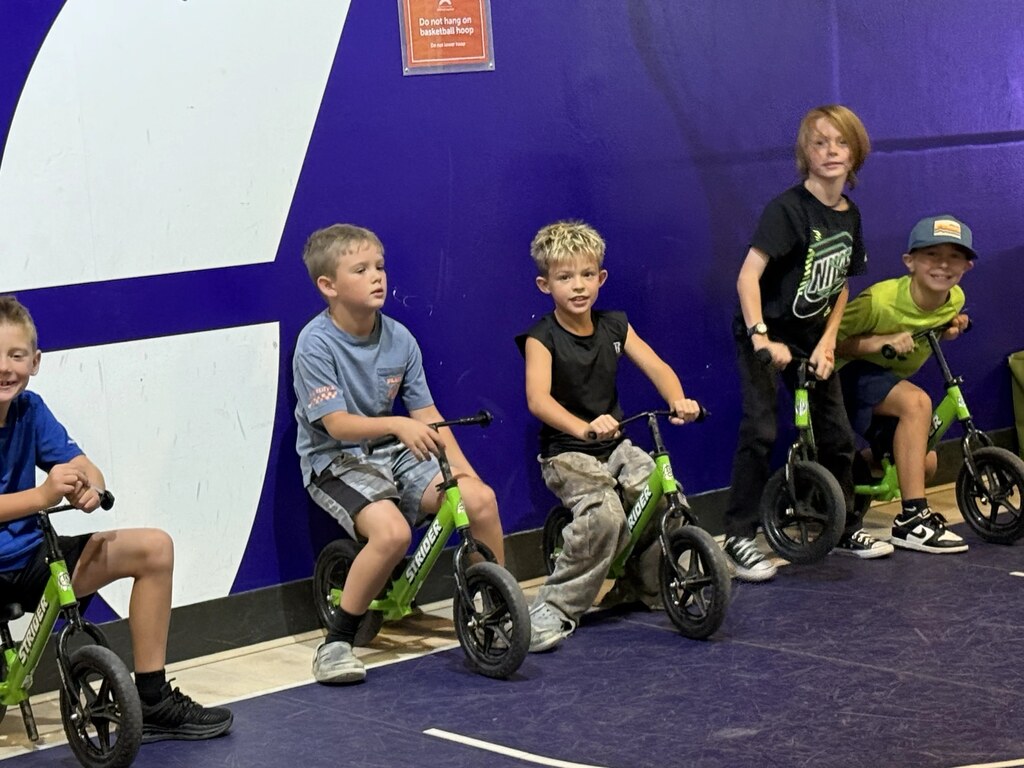 Four elementary students sitting lined up on small green balance bikes indoors against a bright purple wall.