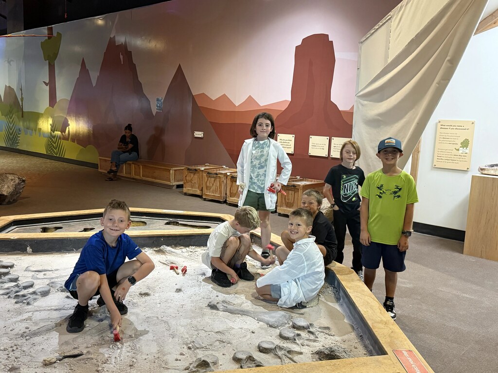 Students posing around a dinosaur dig exhibit pit, with one student wearing a white lab coat.