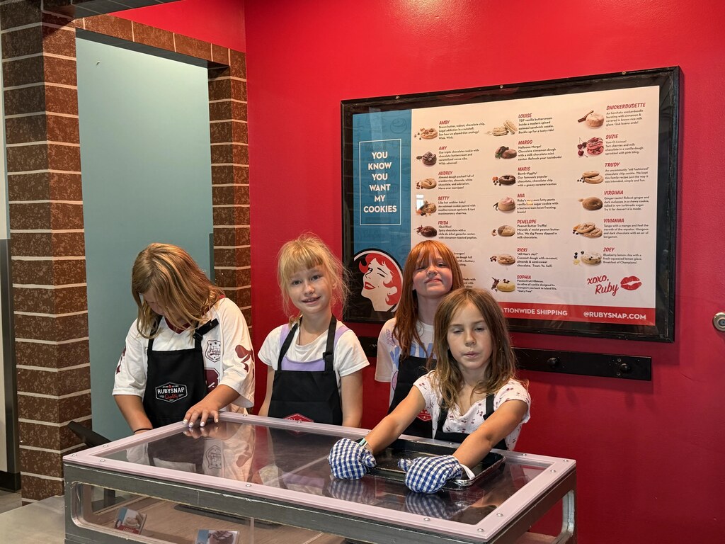 Four students wearing aprons posing behind a display counter at a play bakery exhibit with a large menu board on the wall.