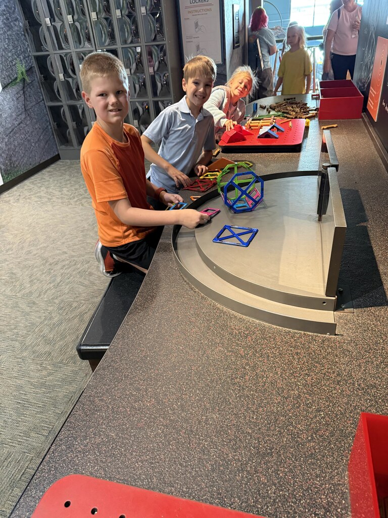 Three students smiling while standing around a tabletop activity station building colorful geometric shapes with magnetic tiles.