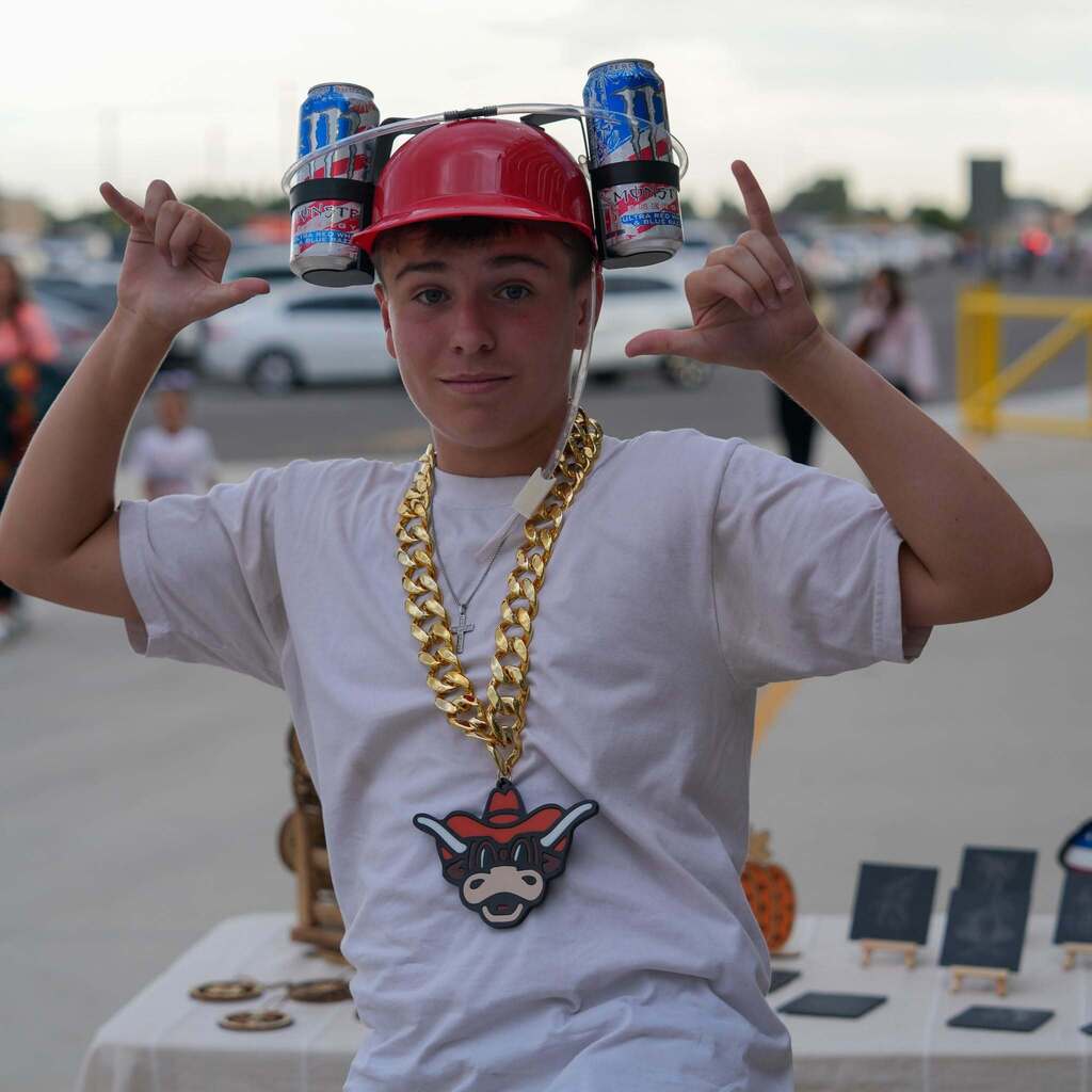 A high school student smiling while wearing a red hard hat drink holder, oversized gold chains, and a mascot medallion.