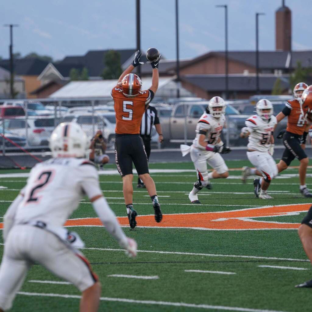 High school football player in an orange jersey leaping high in the air to catch a pass during a game.