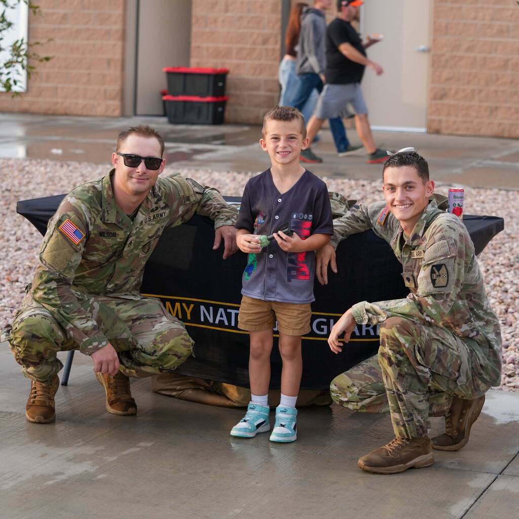 Young boy smiling between two U.S. Army National Guard recruiters crouching at an informational table outdoors.