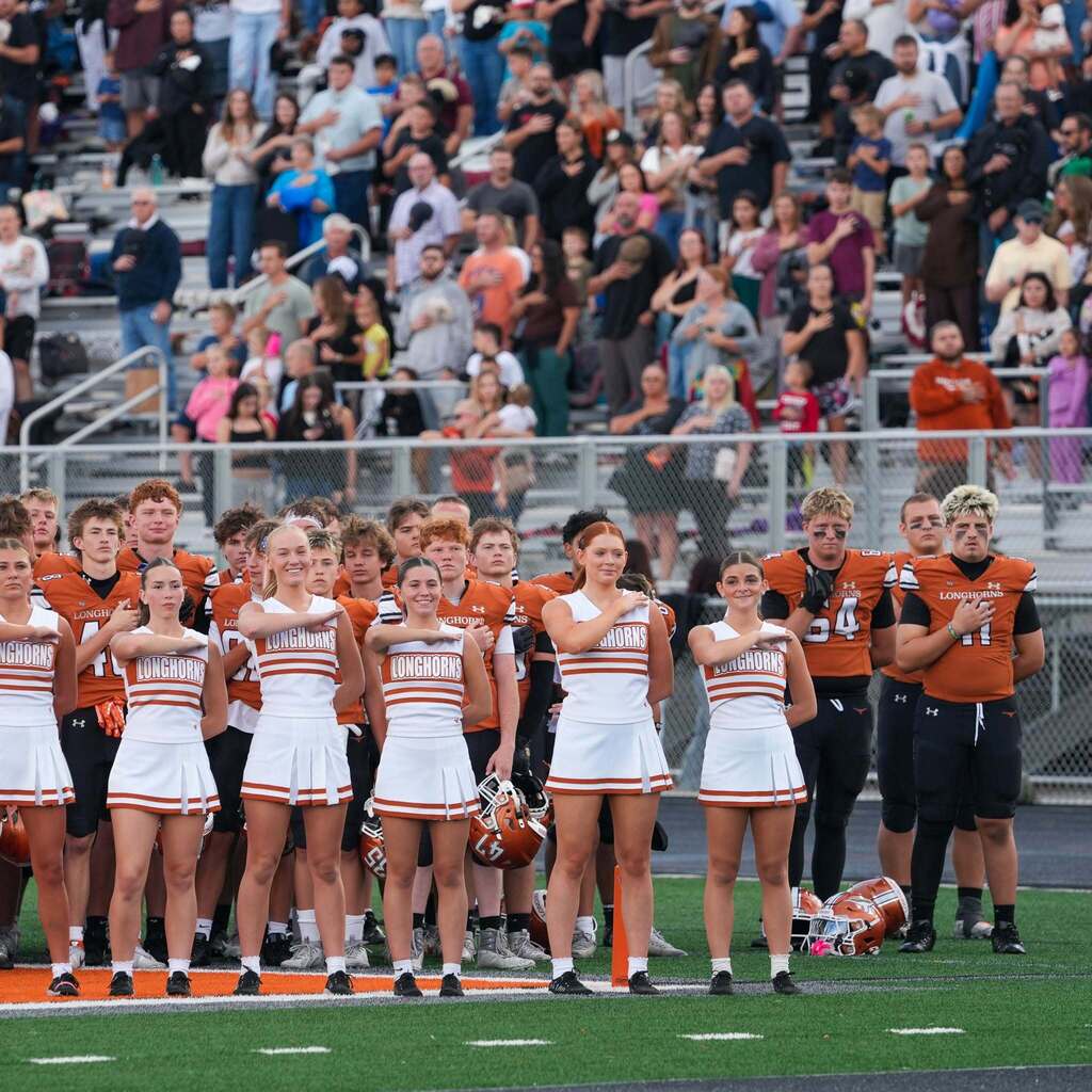 Cheerleaders standing in line in front of football players on the field while fans stand in the bleachers.