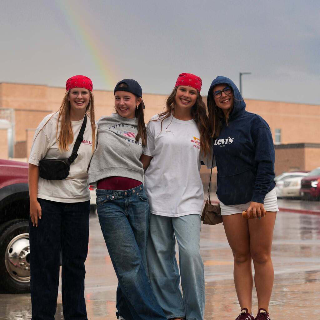 Four high school students posing together outdoors under a rainbow after rain, wearing bandanas and casual gear.