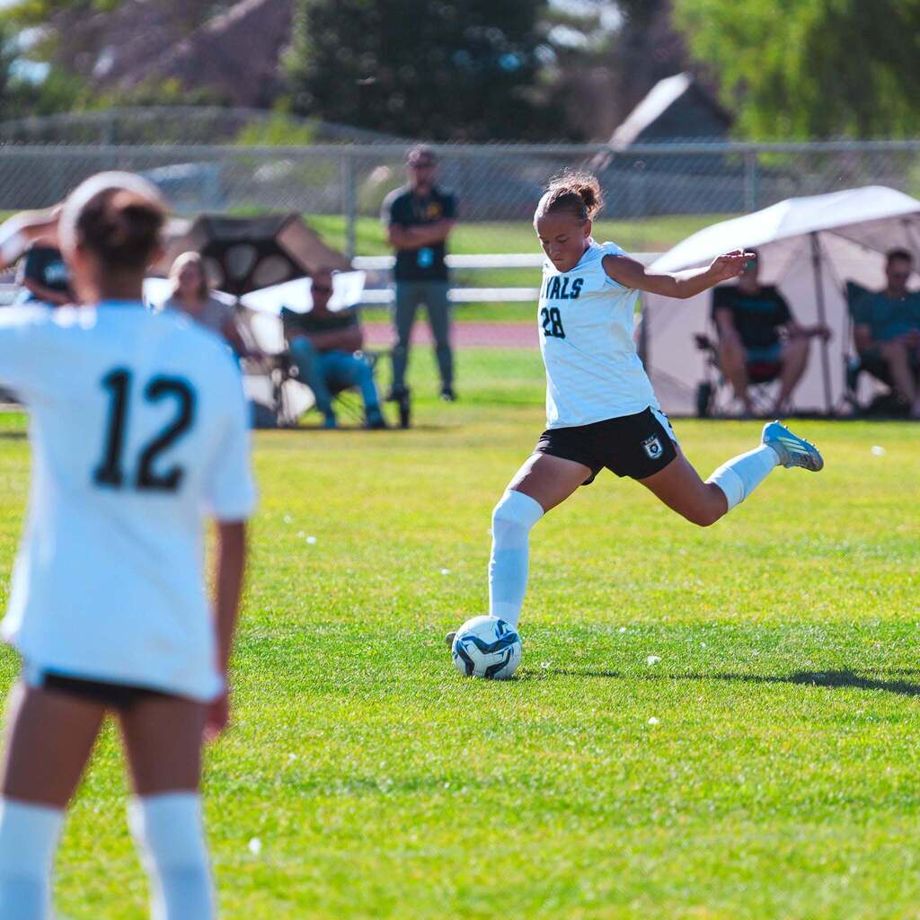 A player in a white soccer jersey winds up to strike a long kick across the grass field during a high school match.