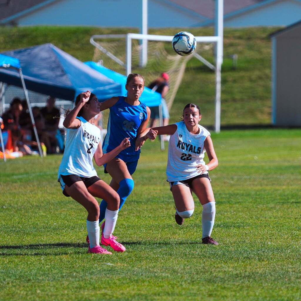 Three soccer players jump into the air together as they contest a high header during a bright game day.
