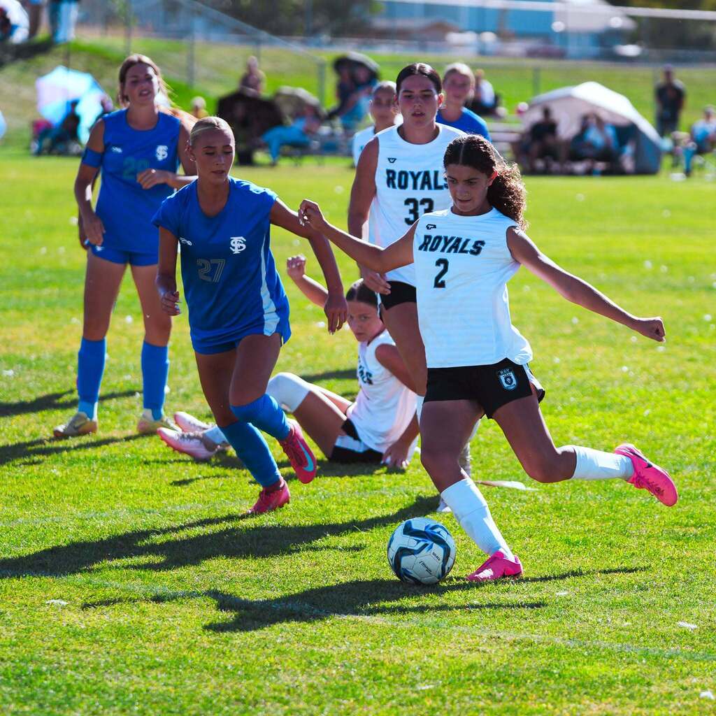 A soccer player in a white jersey advances the ball toward the goal while avoiding a defender in blue.