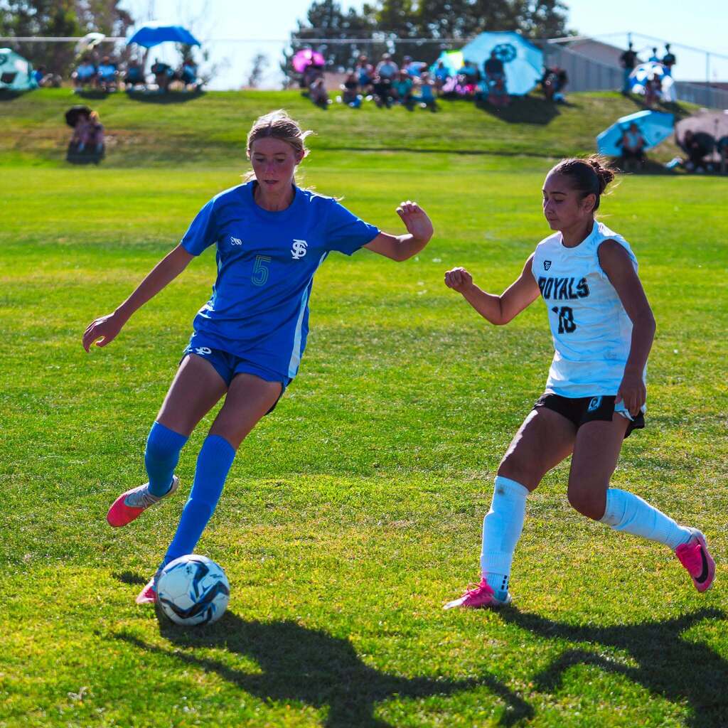 A high school soccer player in a blue uniform dribbles the ball past an opposing defender in a white jersey during a sunny afternoon match.