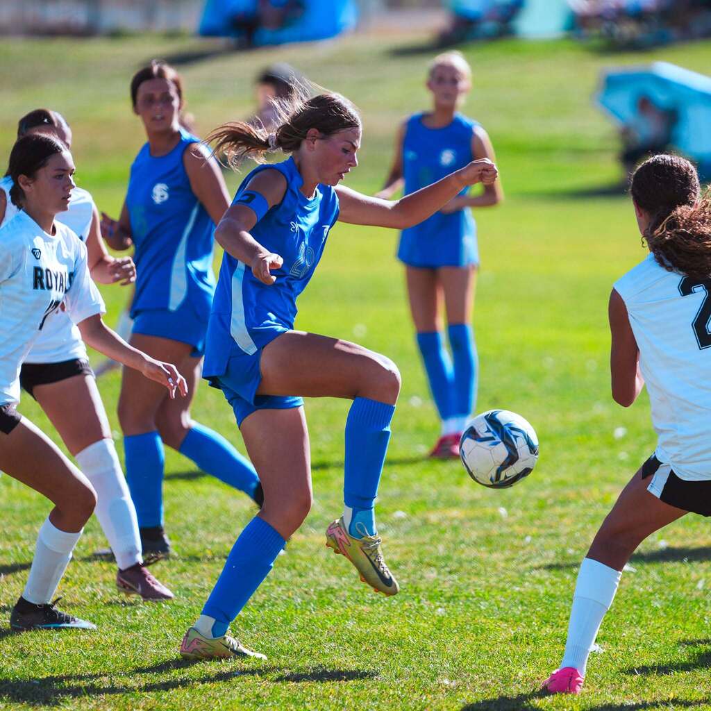 A high school soccer player in blue leaps to control an incoming ball with her knee while surrounded by opposing players.