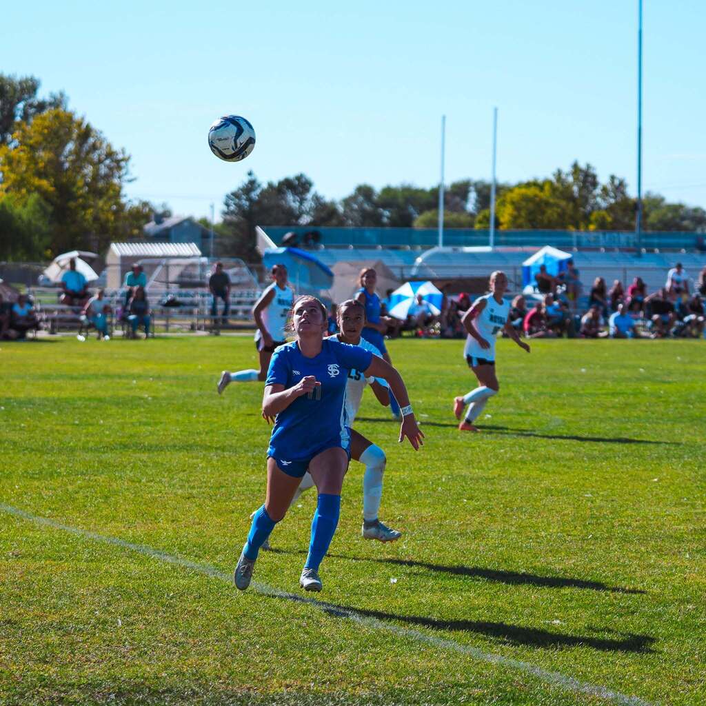 A soccer player in a blue jersey tracks a high ball in the air as defenders close in behind her.