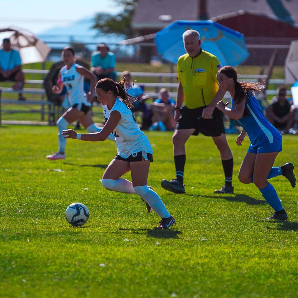 A student-athlete in a white soccer jersey maneuvers the ball downfield, with a blue-jersey defender and referee in hot pursuit.