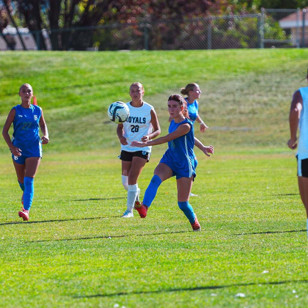 A high school soccer player in a blue uniform tracks the ball mid-air during gameplay on a sunny athletic field.