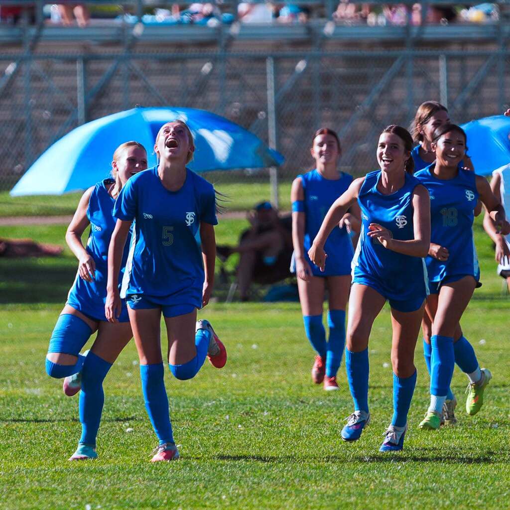 Teammates in blue soccer uniforms sprint across the grass field together, shouting and celebrating a goal.