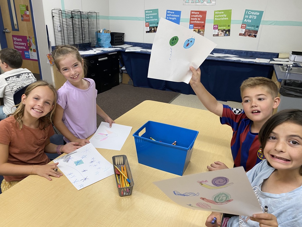 Four 2nd grade Plain City Elementary students standing together in the art classroom, smiling proudly while holding up their colorful, hand-drawn art projects for the camera.