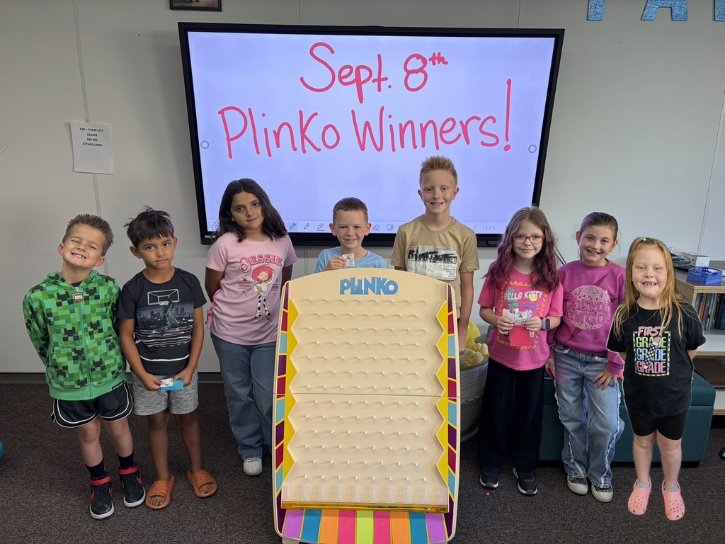 Plain City Elementary students standing together behind a wooden Plinko game board in the school hallway, smiling and holding up their rewarded PAW tickets.