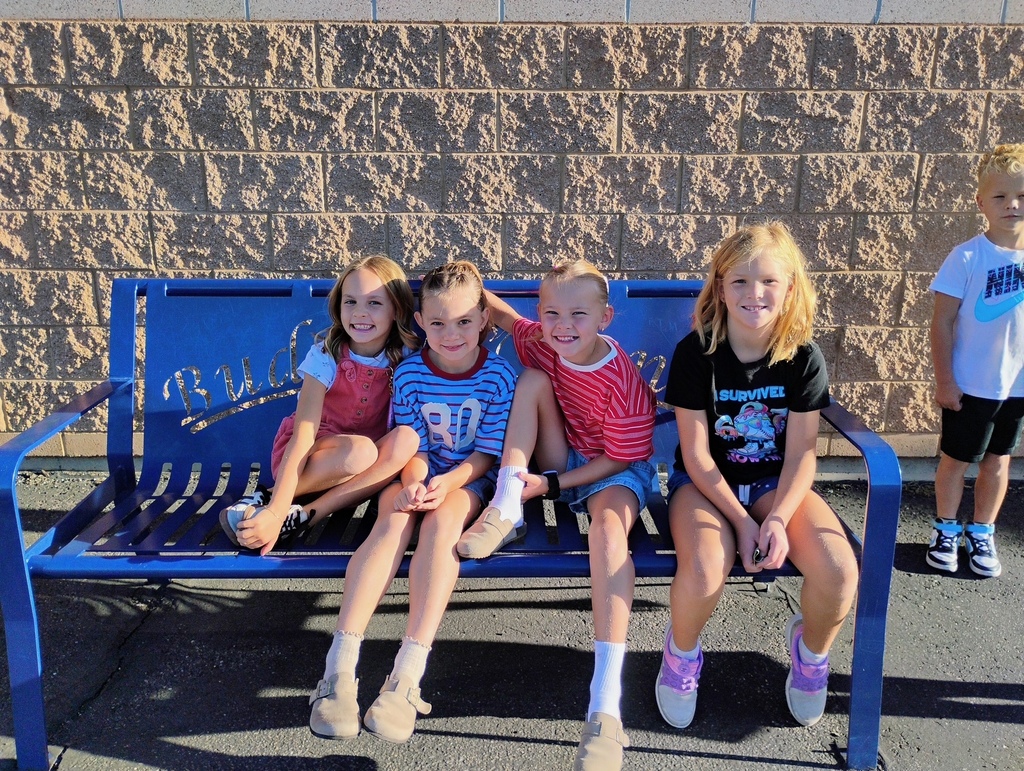 Four young second-grade girls sitting side-by-side on a blue metal buddy bench on the school playground. They are all smiling warmly at the camera, wearing casual school clothes, with the outdoor play area and school building visible in the background.