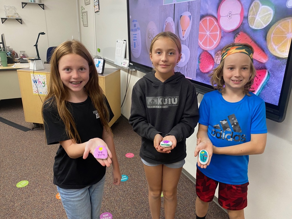 4th-grade students at Plain City Elementary smiles while holding up a freshly painted rock, with colorful paint pens and other decorated rocks visible on the classroom table in front of them.