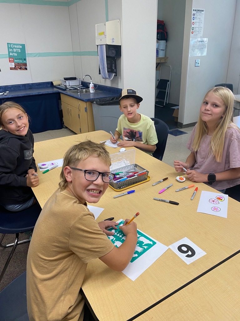 4th-grade students at Plain City Elementary smiles while holding up a freshly painted rock, with colorful paint pens and other decorated rocks visible on the classroom table in front of them.
