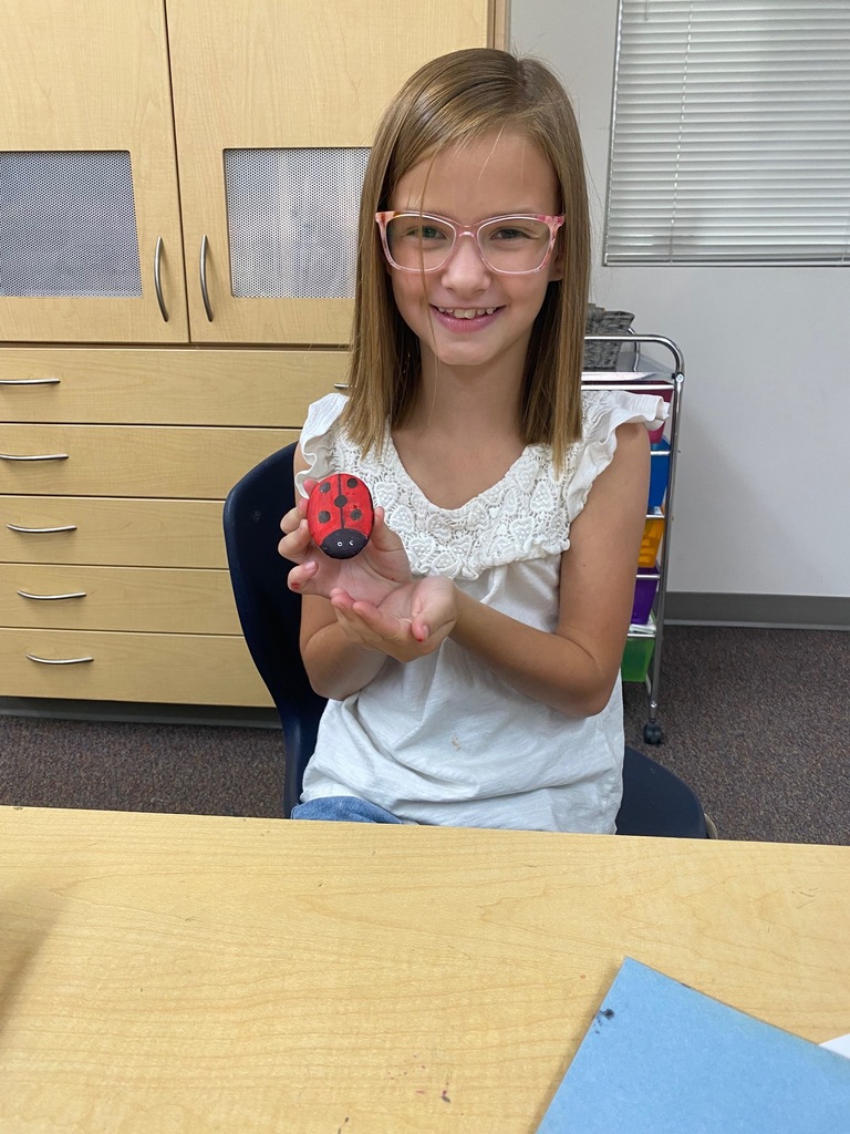 4th-grade students at Plain City Elementary smiles while holding up a freshly painted rock, with colorful paint pens and other decorated rocks visible on the classroom table in front of them.