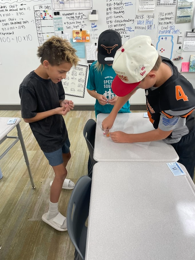 Three boys work together on a team-building activity at a desk in front of a classroom whiteboard. Visible text on the whiteboard includes "IXL codes", "homework is due Friday, Sept 11th", "Picture Day is Tues, Sept 15th", "Math", "Reading", "Lunch", "Week goal", "Voice level", "Reward", "PLC points", "Big Reward", "Goal 40", "Root beer floats", "Looks like decimal shifts", "exponent", and "100 = 10 x 10". Clothing text includes "Haven Bay SPEECH SQUAD", "SAN FRANCISCO", and "85".