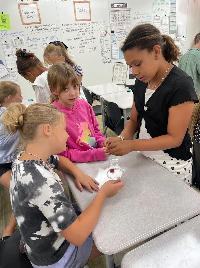 Three female students collaborate on a cup-and-paperclip challenge at a desk with other classmates nearby. Visible text on the background whiteboard includes "Language Arts", "Math", "I can identify text structures of informational passages", "Thoreau was likely feeling mad", "I can multiply & divide by powers of 10", "multiply - numbers shift", "divide - numbers shift", "exponent", "100 = 10 x 10", "fast finisher", and "SEPTEMBER".