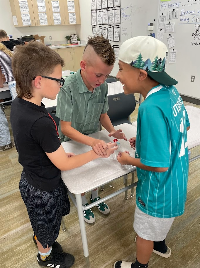 Three male students leaning over a desk while working together to complete a paperclip challenge with a plastic cup and gummy candy. Visible text on clothing and classroom walls includes "UTAH", "fast finisher", "Thoreau was feeling mad", "upset", "overwhelmed", "stressed", and "frustrated".