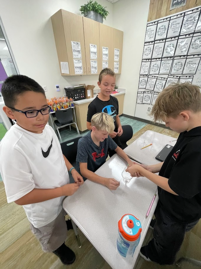 Four boys surrounding a desk conducting a group challenge with a plastic cup, string, and pencils. Visible text in the classroom includes student work displays, a water bottle labeled "Jerics", and clothing logos including "Nike" and "Champion".