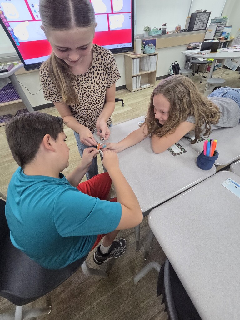 Three students work together on a desk activity in front of a digital display board.