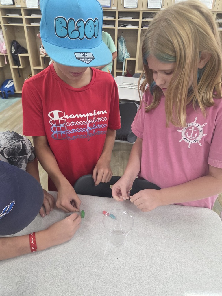 Three young students gather around a desk attempting to rescue a gummy worm under a plastic cup using paperclips without touching it directly. Visible text includes "BL101", "SNAPBACK", "Champion", "ANNAPOLIS MARYLAND", and a red wristband with text.
