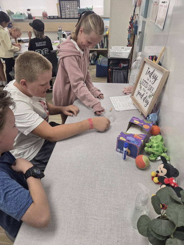 Students gathered at a table working on a hands-on activity near classroom decorations and a framed sign. Visible text includes "VIP TABLE", "BARKELEY", "today is a GOOD DAY for a good day", "Math", and various classroom schedules and charts on the wall.