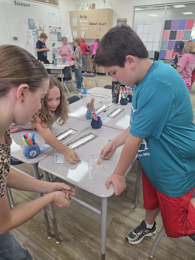 Three students work together at a desk on a science STEM activity involving paperclips, a plastic cup, and a gummy worm, while other classmates work in the background. Visible text on signs and clothing includes "Science", "SOAR", "Help", "Shout Outs", "Est. 2025"