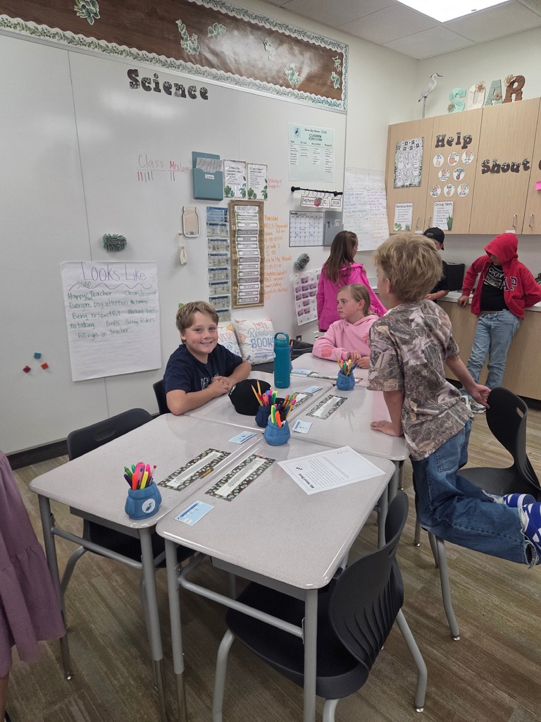 A classroom scene showing elementary students sitting around grouped desks during a group activity. Visible text on the classroom walls and boards includes "Science", "SOAR", "Help", "Shout", "Class Rules", "Looks Like", "Happy Teacher", "Everyone pay attention", "Listen to perfect", "No talking", "Clean floor", "All eyes on teacher", "September 2026", and "Love Reading BOOKS".