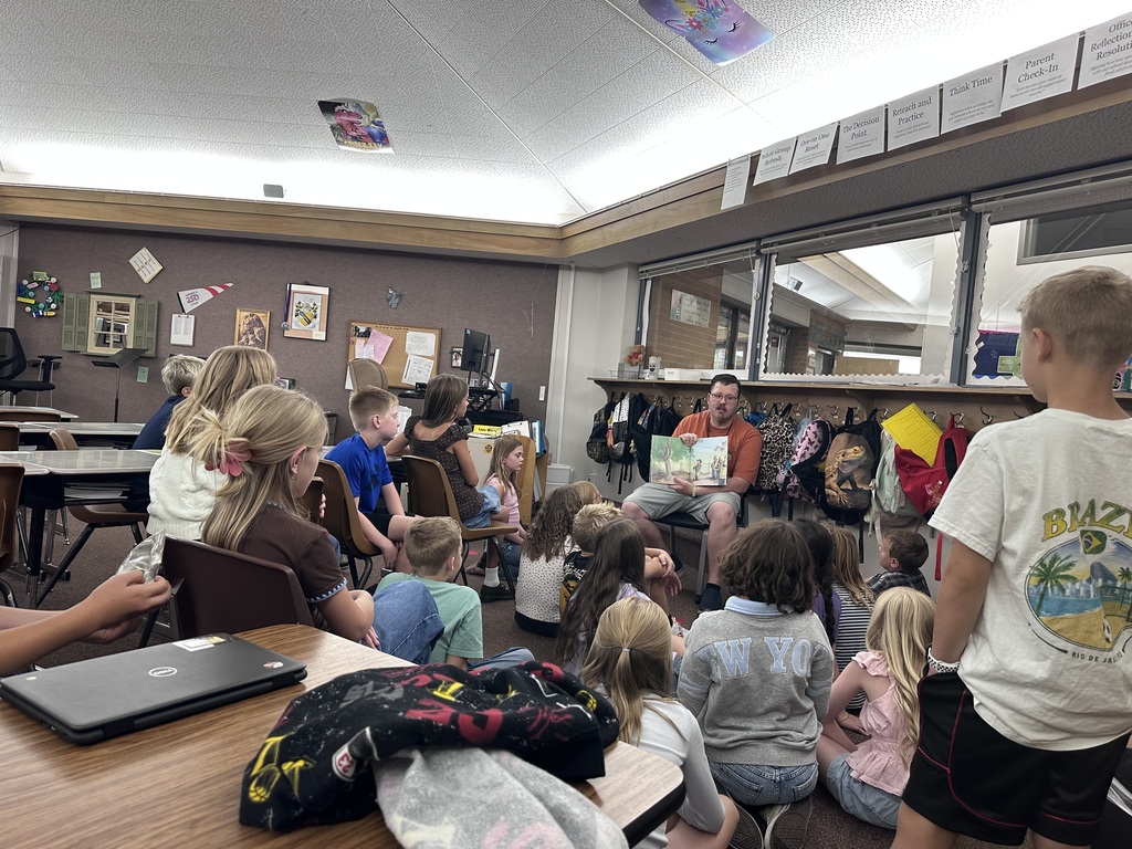 Teacher Mr. O sitting in front of his 5th-grade classroom, holding open a picture book to read aloud while students sit attentively on the carpet and at desks listening.