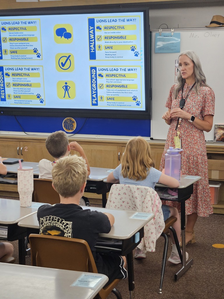 Lomond View Elementary Principal standing at the front of a classroom speaking to seated students, next to a large digital screen displaying "Lions Lead the Way!" expectations for being respectful, responsible, and safe.