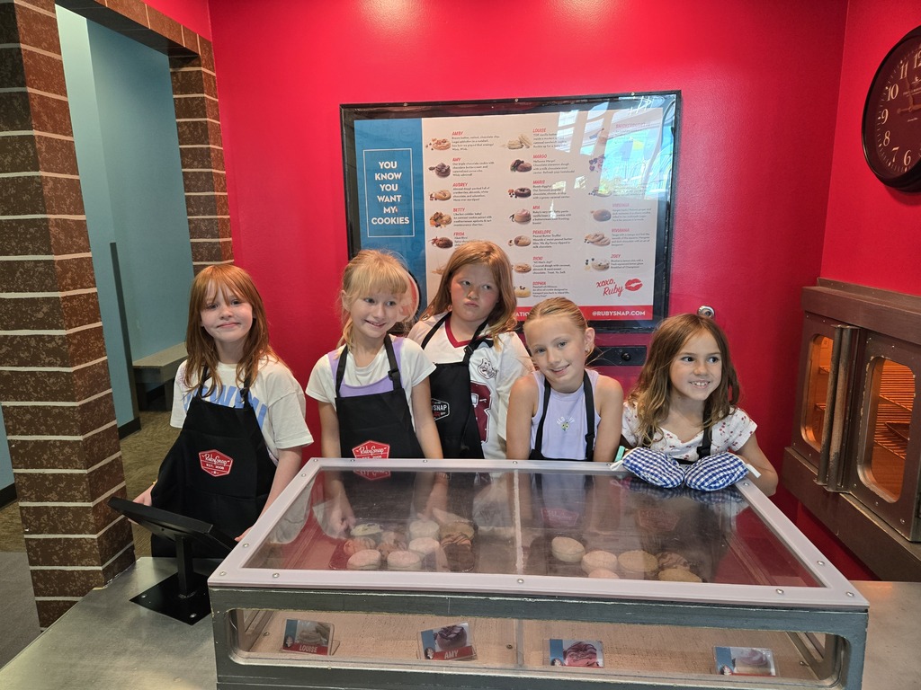  Five girls wearing aprons and oven mitts posing behind a display case at a pretend bakery exhibit.