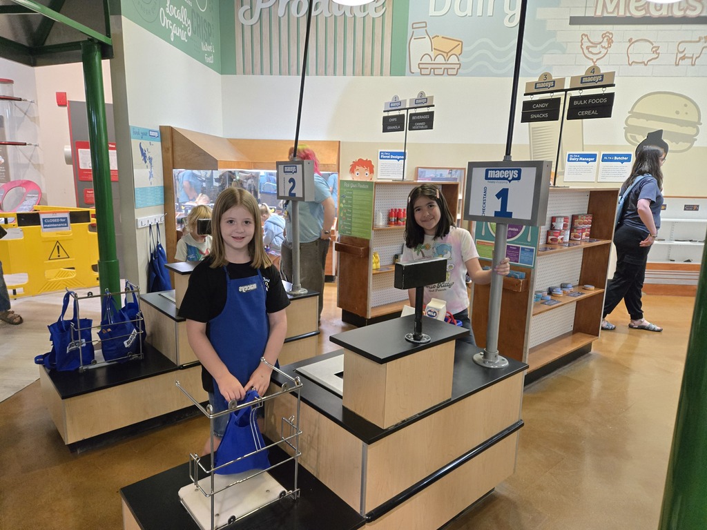Two students posing behind checkout counters at a play grocery store exhibit.