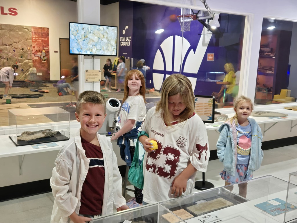 A group of students exploring dynamic science exhibits in a museum gallery, with one student wearing a white lab coat.