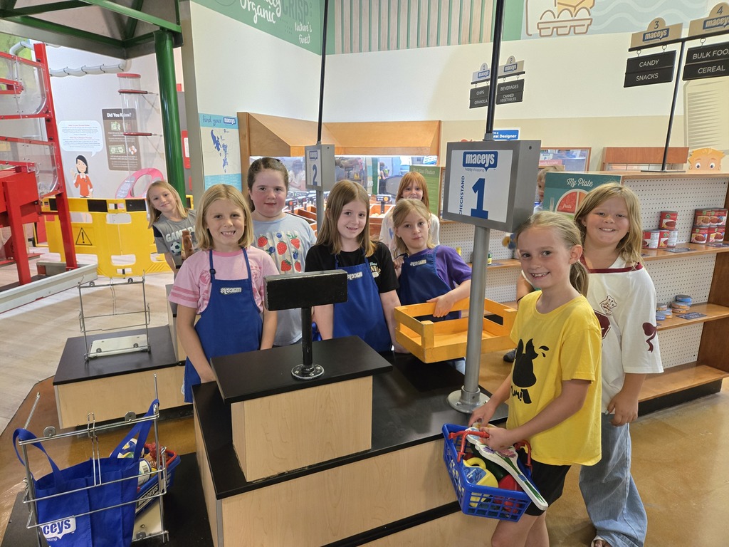 A group of 3rd-grade students smiling behind a pretend grocery store checkstand wearing blue aprons and holding shopping baskets.