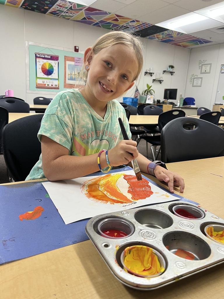 second grade student smiling while paining with orange yellow and red on a  drawn pumpkin