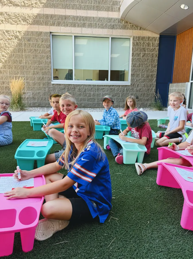 A group of young elementary school students sitting outside on green artificial turf, working on papers placed on colorful plastic lap desks. A blonde girl in the foreground wears a blue sports jersey and smiles at the camera. Other children around her are writing or drawing with markers.