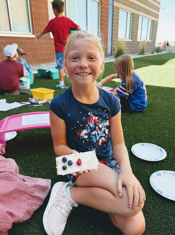 A young girl with blonde hair kneeling on artificial turf outdoors, smiling at the camera while holding up a cracker topped with white spread, four blueberries, and one raspberry to resemble a patriotic flag craft. Other students and plastic lap desks are visible in the background.
