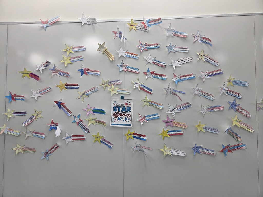 A classroom whiteboard covered with numerous paper shooting star crafts created by students, painted in red, white, and blue designs with written messages. In the center, a printed sign reads "Every STAR can make a difference" surrounded by small star illustrations.
