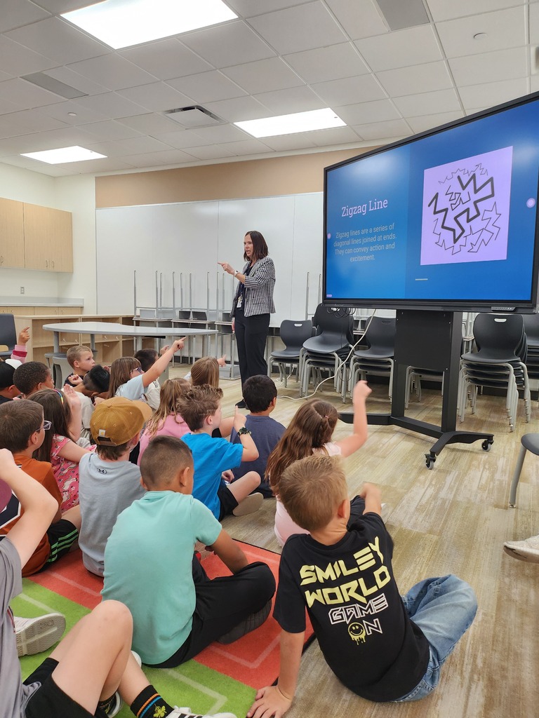 A teacher stands at the front of a classroom presenting a lesson on a large screen displaying the title "Zigzag Line" along with text explaining "Zigzag lines are a series of diagonal lines joined at ends. They can convey action and excitement." A group of young students sits on the carpet listening, with one boy in the foreground wearing a black shirt that reads "SMILEY WORLD GAME ON."